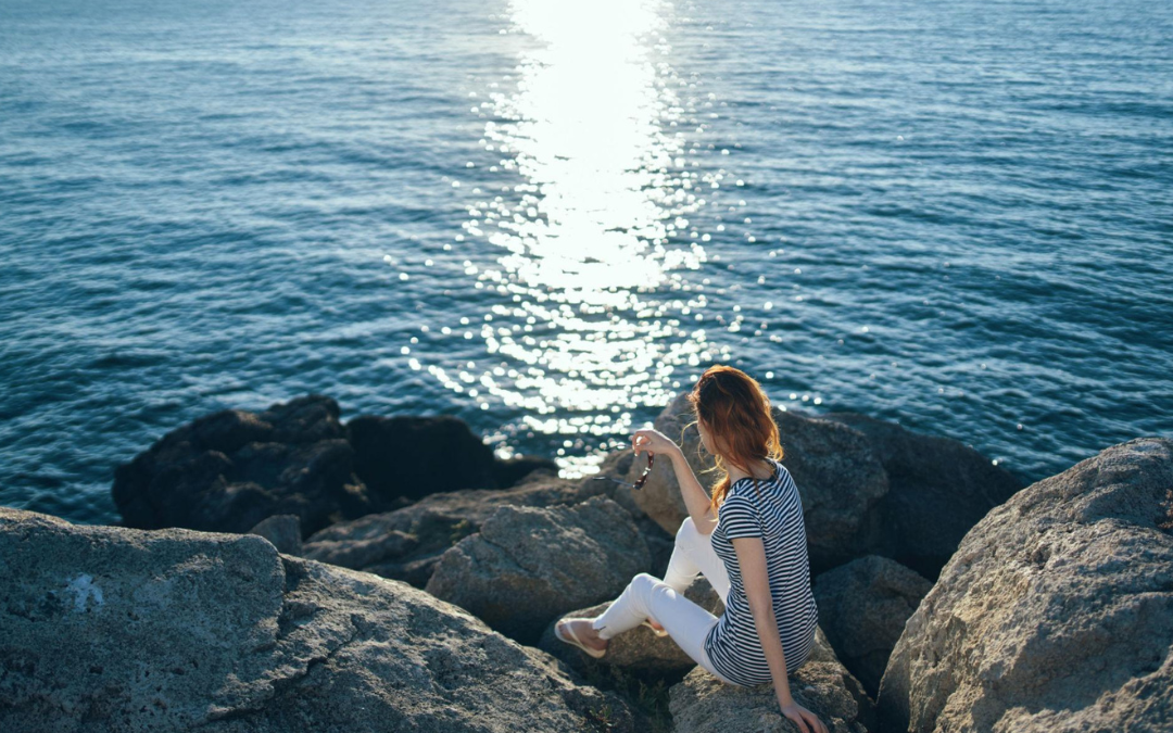 Woman meditating by the ocean at sunrise with healing crystals and a travel journal during a spiritual healing journey.