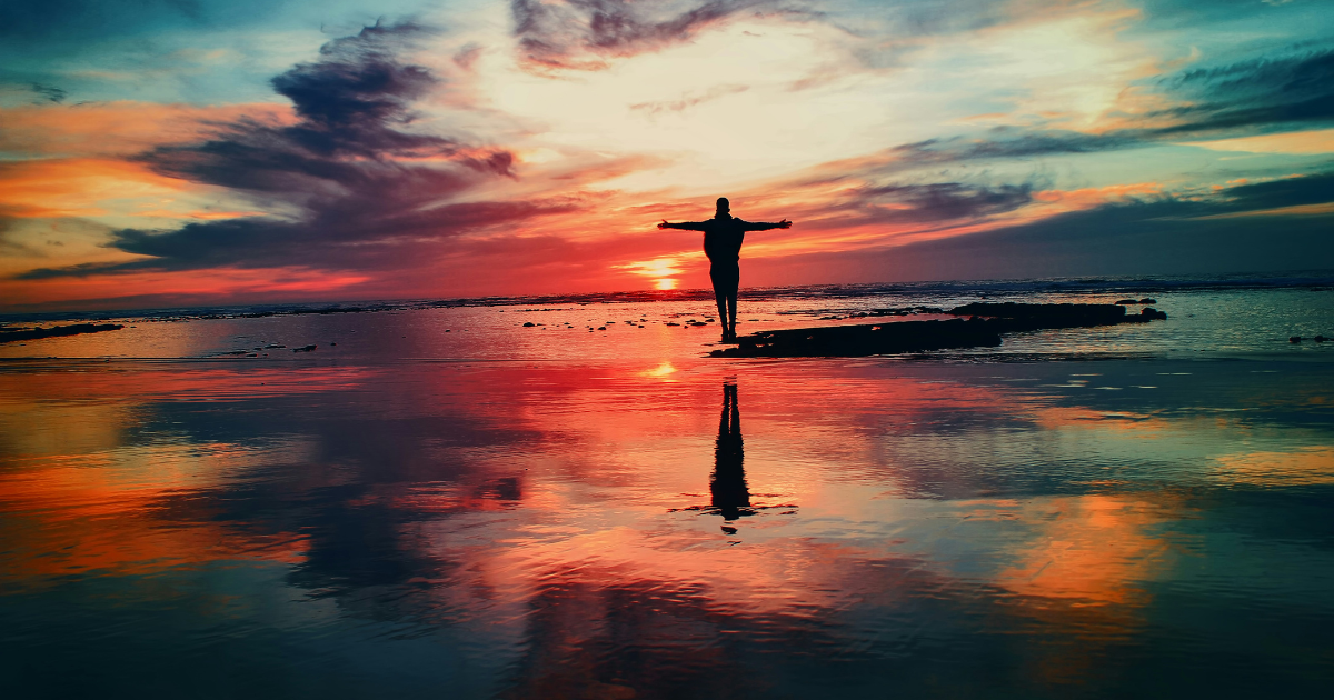 The Independence We Forget: Freedom From the Need to Be Someone Else — a person standing with arms outstretched beside the sea at sunset, symbolizing freedom and self-trust