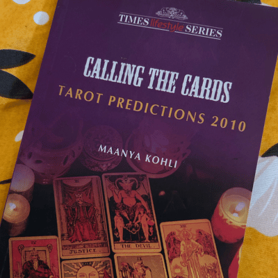 Book cover of 'Calling the Cards: Tarot Predictions 2010' by Maanya Kohli, part of the Times Lifestyle Series, resting on a yellow floral fabric. The cover shows tarot cards (Justice, The Devil, Temperance, The Emperor, The Sun, The Hermit) arranged among candles